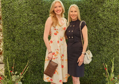 Two women stand smiling in front of a green hedge wall with a neon “AR KIDS READ” sign. One wears a floral dress and white heels, the other a navy dress and black heels. Decorative plants and watering cans are on the floor.