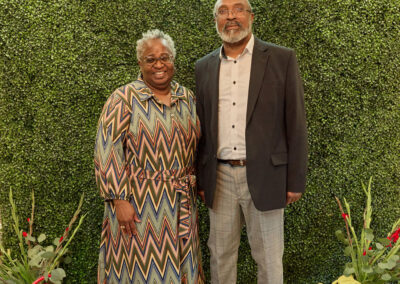 Two adults stand smiling in front of a green leafy backdrop with a neon sign that reads AR KIDS READ. They are dressed formally, with flowers and watering cans on either side of the rug beneath them.