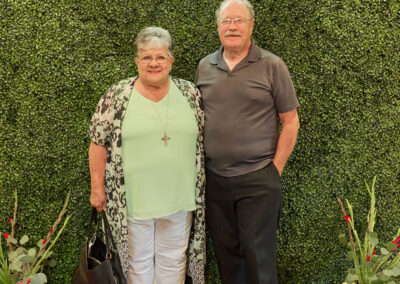 An older woman and man stand smiling in front of a leafy green wall with a neon sign that reads AR Kids Read. They are surrounded by decorative plants and colorful watering cans on a patterned rug.