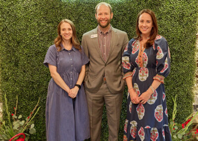 Three adults stand smiling in front of a green leafy wall with a neon sign that reads AR KIDS READ. Two women in dresses flank a man in a suit. Decorative plants and a red watering can are on the floor nearby.