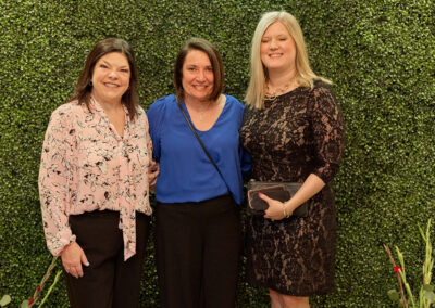 Three women stand smiling in front of a green leafy backdrop with a neon sign that reads AR KIDS READ. Two small watering cans and flowers sit on the floor on either side of them.