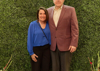 A woman in a blue blouse and black pants stands next to a man in a maroon blazer and gray pants, posing in front of a green leafy backdrop with a neon sign that reads AR Kids Read. Gardening tools and flowers decorate the scene.
