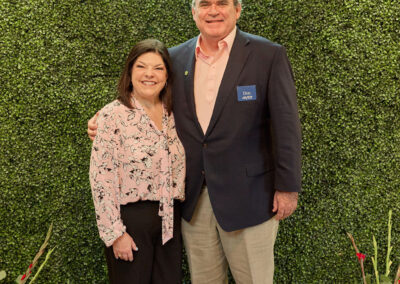 A smiling woman and man stand together in front of a green leafy wall with a neon sign that reads AR KIDS READ. Watering cans and flowers are placed at their feet.