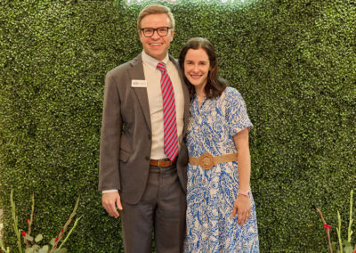 A man in a suit and a woman in a blue and white dress stand smiling in front of a green leafy wall with a neon sign that reads “AR Kids Read.” Watering cans and flowers decorate the bottom of the wall.