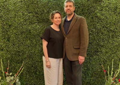 A woman and man stand together, smiling in front of a green leafy backdrop with a neon sign that reads AR KIDS READ. Two small watering cans and plants are placed on the floor at each side of the couple.