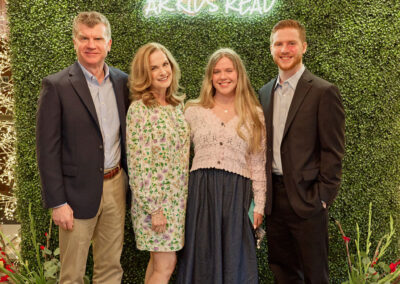 Four people stand smiling in front of a green leafy wall with a neon sign that reads AR KIDS READ. Two men in suits stand on either side of two women in floral dresses. Decorative plants are visible on the floor.