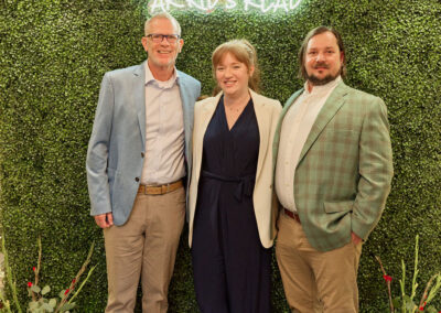 Three people stand smiling in front of a green foliage wall with a neon sign that reads “AR Kids Read.” Two men in blazers flank a woman in a dark jumpsuit and white blazer. Decorative watering cans are on the floor.