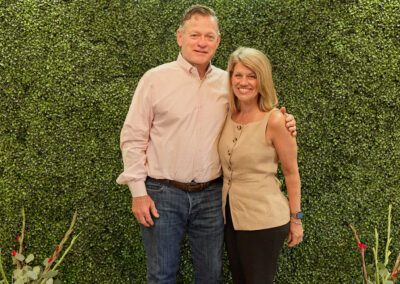A man and woman stand smiling together in front of a green leafy wall with a neon sign that reads AR KIDS READ. Decorative watering cans and plants are on either side of them.