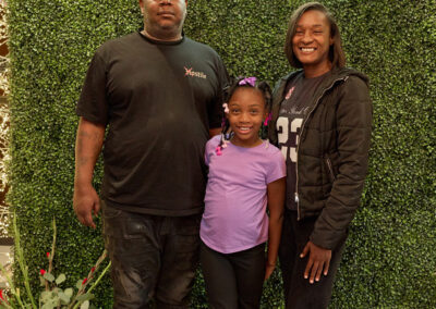 A man, a woman, and a young girl stand smiling in front of a green leafy wall with a neon sign reading AR Kids Read. A pink watering can and flower arrangement are on the floor beside them.