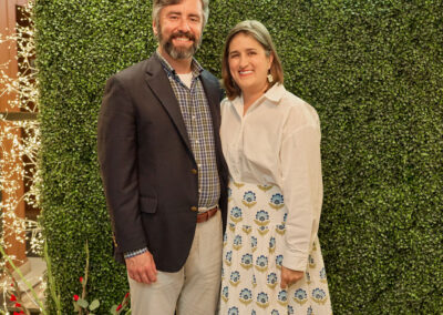 A man and woman stand smiling in front of a faux greenery wall with a neon sign that reads AR KIDS READ. Decorative lights, a red watering can, and flowers are visible to the left.