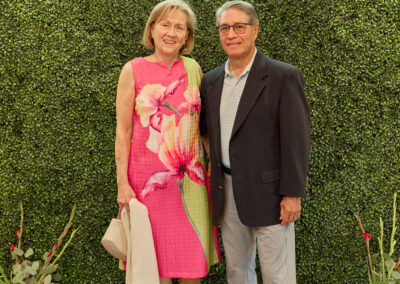 A smiling woman in a pink floral dress and a smiling man in a dark blazer stand together on a patterned rug, in front of a green hedge wall with a neon sign that reads ARKIDSREAD. Watering cans and flowers are on the ground.