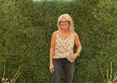 A woman with blonde hair, glasses, and a heart-patterned top stands smiling in front of a green faux-hedge wall with a neon AR KIDS READ sign. She holds a brown bag; watering cans and flowers are on each side.