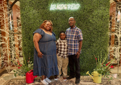 A woman, child, and man stand together smiling in front of a green leafy backdrop with a neon sign reading AR KIDS READ. Decorative lights, red flowers, and yellow watering cans are on the floor beside them.