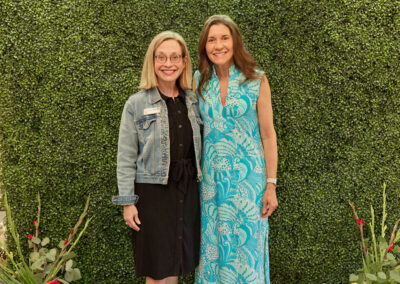 Two women smiling and standing side by side in front of a green leafy wall with a neon sign that says AR KIDS READ. Flowers and colorful watering cans are placed on the floor nearby.