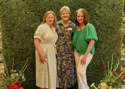 Three women stand smiling in front of a green hedge wall with a neon sign that reads AR KIDS READ. Decorative watering cans and flowers are arranged at the bottom of the backdrop.