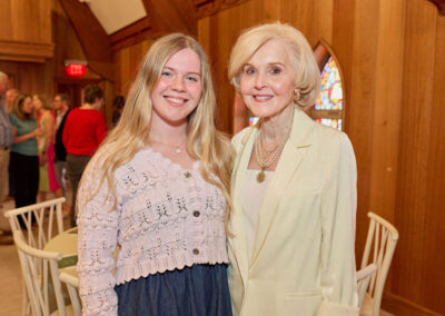 Two women, one younger with long blonde hair and one older with short blonde hair, stand smiling together in a warmly lit room with wooden walls and stained glass windows. People are gathered in the background.