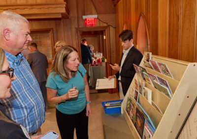A group of people stand by a wooden display rack holding colorful cards or brochures in a wood-paneled room. One woman closely examines the display while others watch or use their phones. A man stands nearby holding a folder.