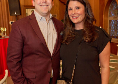 A man in a maroon blazer and a woman in a black dress stand smiling together in a warmly lit wooden room with arched windows and banquet tables.