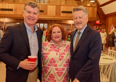 Three adults, two men in suits and a woman in a pink and white floral dress, smile together at an indoor social event with tables and people in the background. One man holds a red cup.