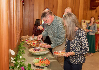 Several people serve themselves food from a buffet table covered with vegetables, dips, and snacks in a wood-paneled room, with others waiting in line and talking in the background.