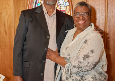 An older man in a black suit and an older woman in glasses and a patterned blouse smile and pose together in front of a wooden wall with a stained glass window.