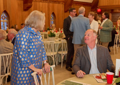 A woman in a blue patterned dress stands talking to a seated man in a brown checked jacket at an indoor event. Other guests mingle in the background around decorated tables. The setting has wooden walls and arched windows.