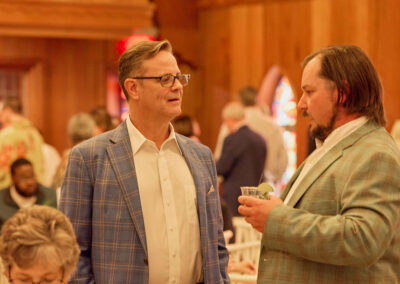 Two men in suits have a conversation at an indoor event with wooden walls. One man holds a drink and they appear engaged in discussion. There are other people seated and standing in the background.
