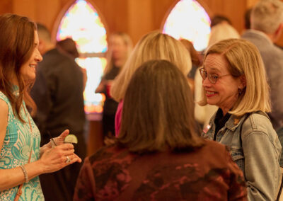 A group of women chat and smile at an indoor social gathering, with stained glass windows in the background creating a warm, inviting atmosphere. One woman holds a drink with a slice of lime.