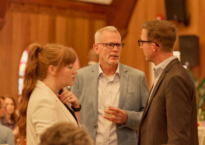 Three people in business attire stand indoors, engaged in conversation. A man in a light blue blazer holds a drink and talks to a woman with red hair and another man in a dark suit. The background features wooden walls and blurred attendees.