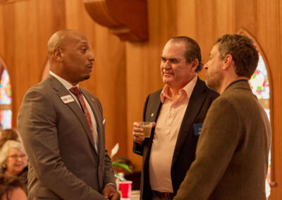 Three men in formal attire are standing and conversing at an indoor event with wooden walls and stained glass windows in the background. One man is holding a drink. Other attendees are seated in the background.
