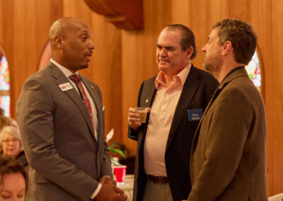 Three men stand and talk at an indoor event with wooden walls and stained glass windows; one man holds a drink and wears a name tag that says Don. Other people are seated in the background.