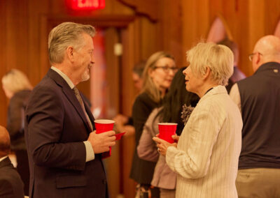 Two older adults in business attire hold red cups and converse at an indoor event, with other people socializing in the background. The room has wooden walls and warm lighting.