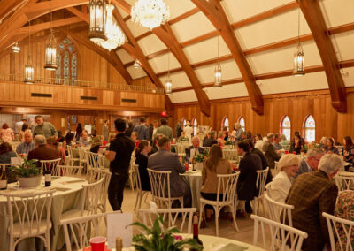 A large group of people sits at round tables covered with tablecloths inside a wooden hall with arched ceilings and chandeliers, attending a formal event or banquet. Stained glass windows line the walls.