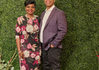 A smiling woman in a floral dress and a man in a plaid blazer stand together in front of a green leafy wall with a neon AR Kids Read sign. There are red and yellow watering cans and plants on the floor nearby.