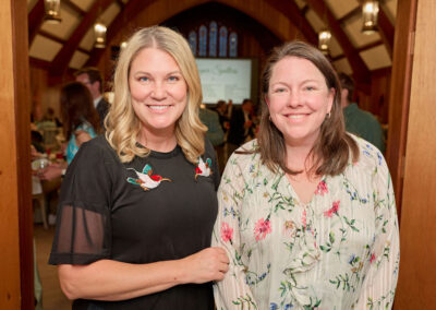 Two women smiling and standing together indoors at an event. One wears a black dress with embroidered birds; the other wears a white floral blouse. People and tables are visible in the background, suggesting a social gathering.