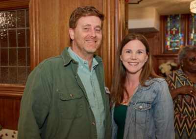 A man in a green jacket and a woman in a denim jacket smile together indoors at a social event. A woman in a patterned dress stands in the background near stained glass windows.