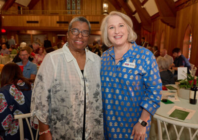 Two women smiling and posing together at an indoor event with wooden walls and arched windows. People are seated at round tables in the background, suggesting a social gathering or banquet.