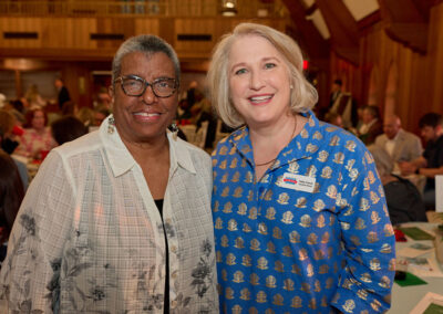 Two women smiling and standing close together at an indoor event, with tables and people in the background. The woman on the left wears glasses and a sheer white shirt, and the woman on the right wears a blue patterned top with a name tag.
