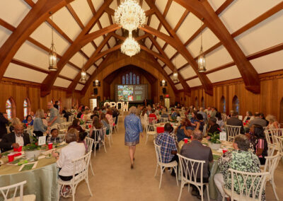 A large gathering of people seated at round tables in a grand hall with wooden beams, chandeliers, and stained glass windows, attending an event with a screen and decorations at the front.