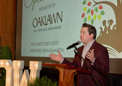 A man in a suit speaks at a wooden podium with a microphone. Behind him is a screen displaying event information, logos, and Wi-Fi details. Large decorative letters and green plants are visible in the background.