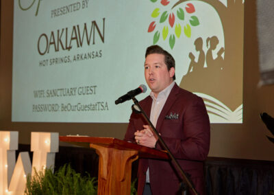 A man in a maroon jacket speaks at a podium with a microphone. Behind him is a projector screen displaying an event presentation with the text OAKLAWN HOT SPRINGS, ARKANSAS and WiFi information.