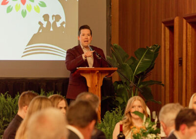 A man in a burgundy jacket speaks at a wooden podium during an indoor event, with an audience seated at tables. A large screen and green plants are visible in the background.
