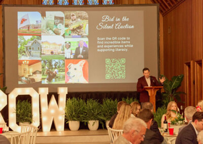 A man speaks at a podium in front of a large screen displaying silent auction info with a QR code. People sit at tables with plants and large illuminated GROW letters in the background.