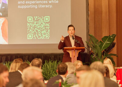 A man in a suit speaks at a podium in front of an audience. Behind him is a projector screen displaying a QR code and text promoting literacy. There are plants and decorations around the stage.