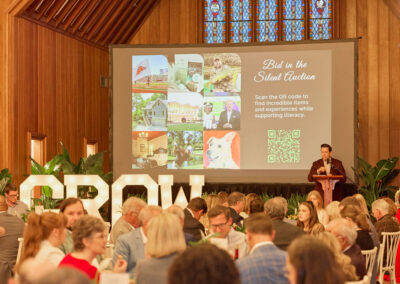 A man speaks at a podium during a formal event in a wood-paneled room. Guests sit at round tables as a large screen displays information about a silent auction supporting literacy. Large illuminated “GROW” letters are visible.