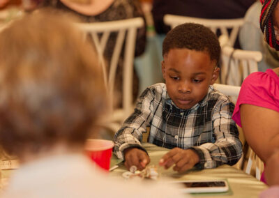 A young boy in a plaid shirt sits at a table, focused on items in front of him. He is surrounded by people, with one person in a pink shirt beside him and a red cup and phone on the table.