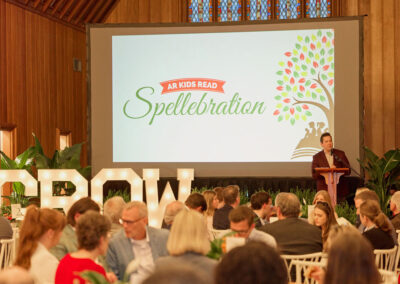 A speaker stands at a podium in a wood-paneled room, addressing seated guests. A large screen behind him displays AR Kids Read Spellebration with a tree graphic. Large lit letters spell GROW.