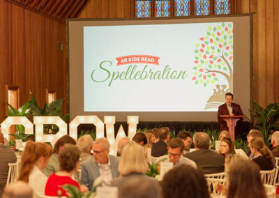 A speaker stands at a podium beside a large screen displaying “AR Kids Read Spellebration” at a formal event attended by seated guests in a wood-paneled room with large “GROW” letters and plants as decor.