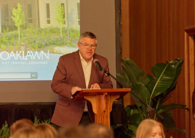 A man in a maroon blazer speaks at a wooden podium with a microphone. Behind him is a projection screen displaying Oaklawn Hot Springs, Arkansas and green plants are nearby. An audience is seated in front.