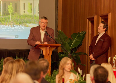 A man speaks at a podium during an indoor event, while another man stands nearby smiling. People are seated in the foreground and a projector screen displays OAKLAWN HOT SPRINGS, ARKANSAS.